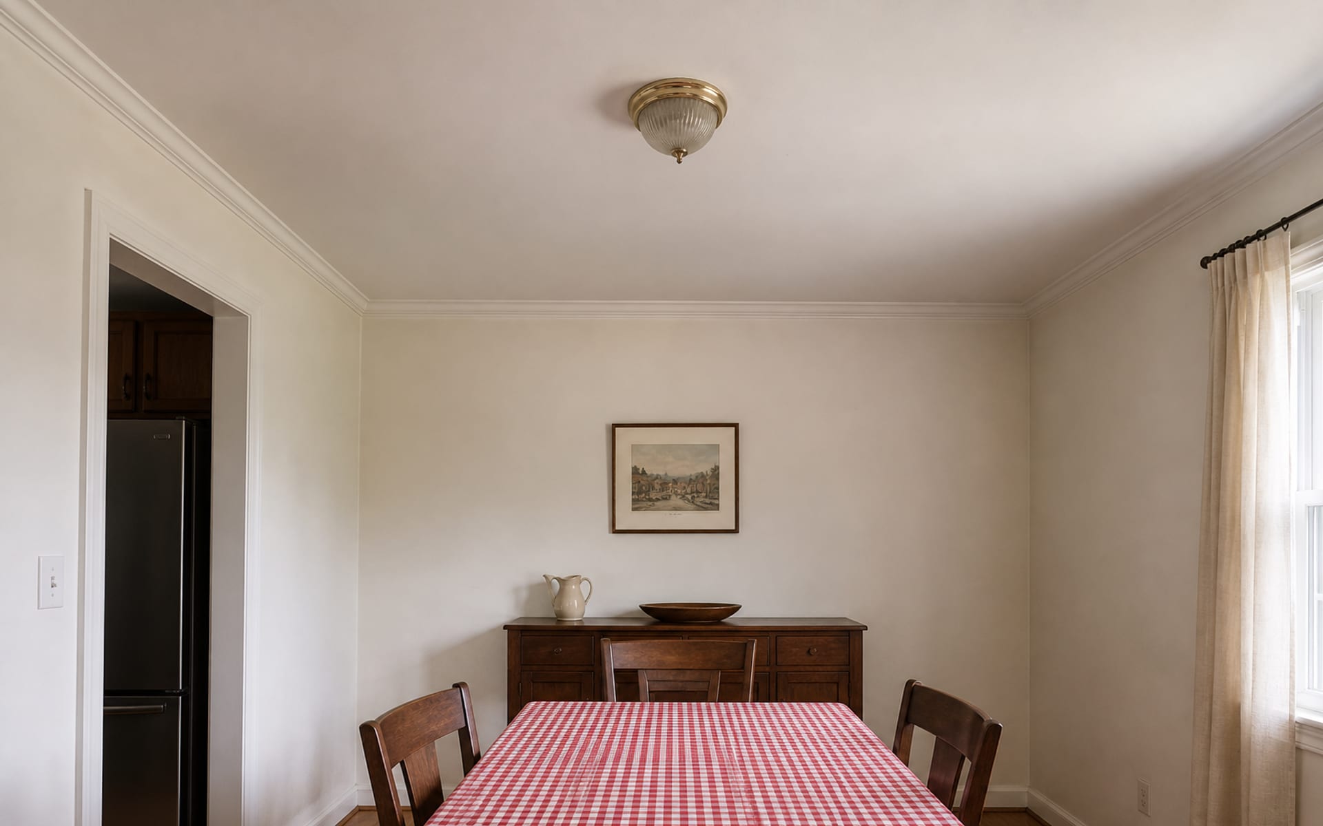 Small dining room with a flat builder-white ceiling, flush dome light, scuffed walls, and a vinyl gingham tablecloth.
