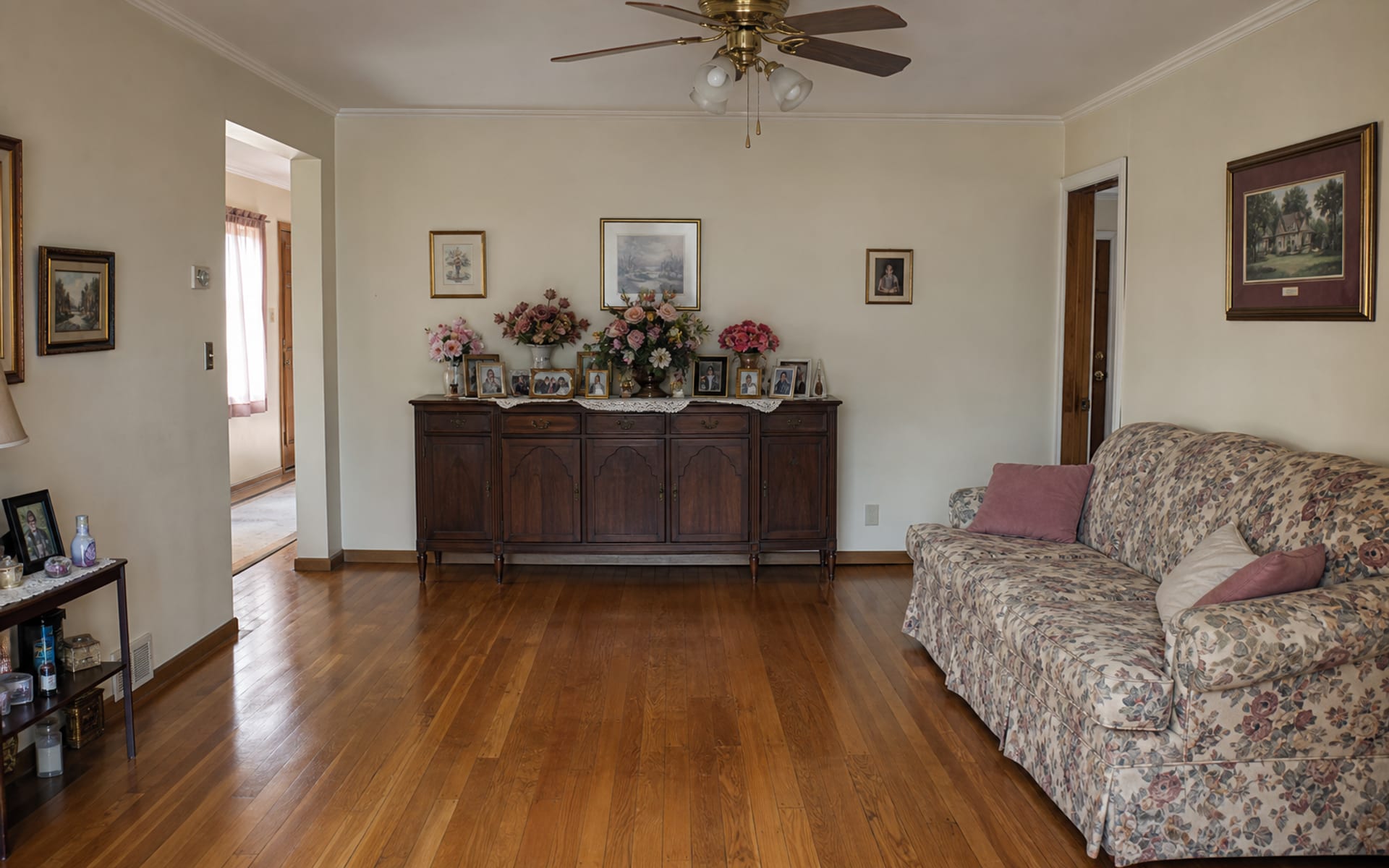 Living room with a cluttered antique sideboard, a dated floral sofa, scuffed beige walls, and harsh overhead light.